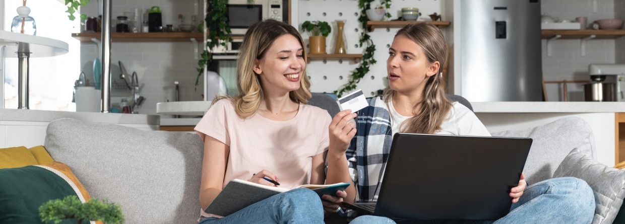 Twee jonge vrouwen zitten op de bank en gebruiken een laptop en een creditcard om online te winkelen